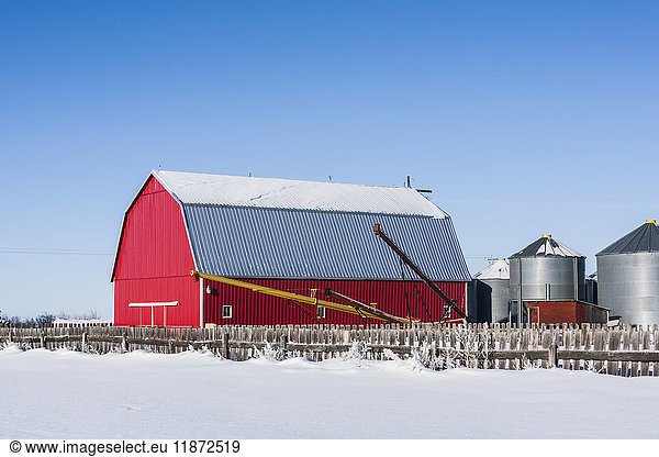 'A bright red barn with a snowy field and wooden fence in the foreground  under a blue sky; Alberta  Canada'