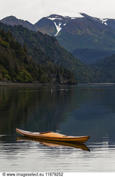 'A kayak sits on the tranquil water with a view of forests and a mountain range in the background  Kachemak Bay State Park; Alaska  United States of America'