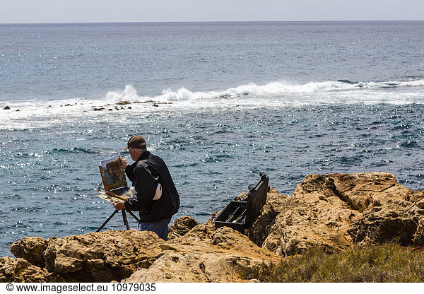 'A man on the seashore painting a coastal scene from an easel; Poipu  Kauai  Hawaii  United States of America'