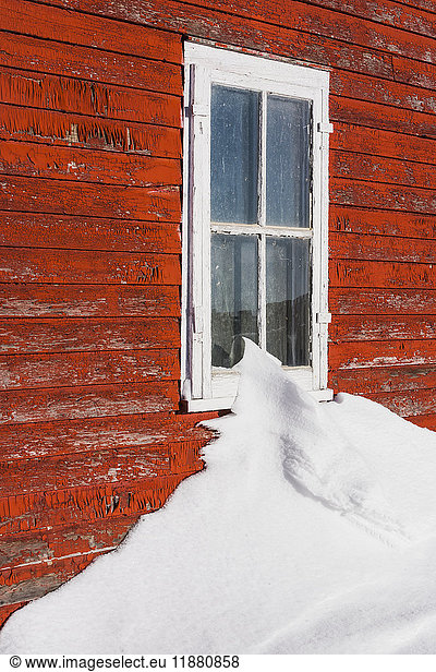 'A snow drift touching the window along a building with a red painted wooden facade that is worn and weathered; Orion  Alberta  Canada'