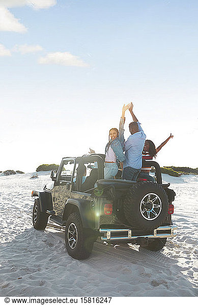 Übermütige junge Freunde  die mit erhobenen Armen im Jeep am Strand jubeln und einen Road Trip genießen