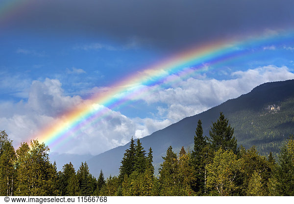 'Colourful rainbow across the sky with mountain side  storm clouds and blue sky in the background; Revelstoke  British Columbia  Canada'
