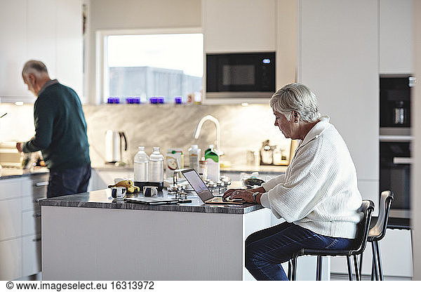 Ältere Frau benutzt Laptop  während ein Mann in der Küche Essen zubereitet