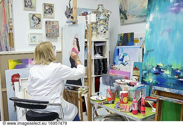 Ältere Frau mit Brille und weißer Schürze steht im Atelier und arbeitet an einem Gemälde