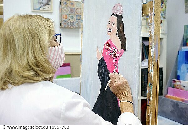 Ältere Frau mit Brille und weißer Schürze steht im Atelier und arbeitet an einem Gemälde