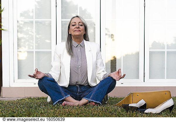 Ältere Geschäftsfrau meditiert im Lotussitz im Gras vor der Bürotür