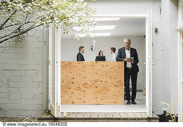 Älterer Geschäftsmann im Büro stehend  mit digitalem Tablet  Menschen arbeiten im Hintergrund