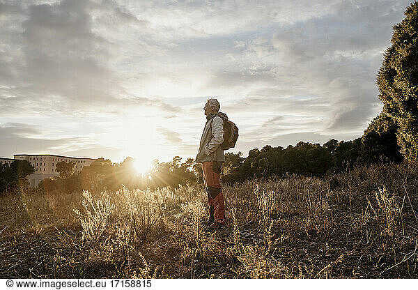 Älterer männlicher Wanderer mit den Händen in den Taschen steht gegen den bewölkten Himmel in der Landschaft bei Sonnenuntergang