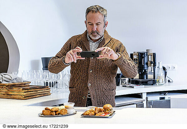 Älterer Mann fotografiert Fast Food mit seinem Smartphone  während er in der Küche zu Hause steht