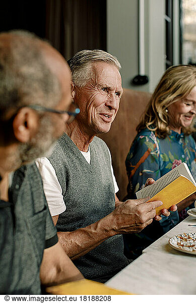 Älterer Mann hält ein Buch in der Hand und unterhält sich mit männlichen und weiblichen Freunden in einem Café