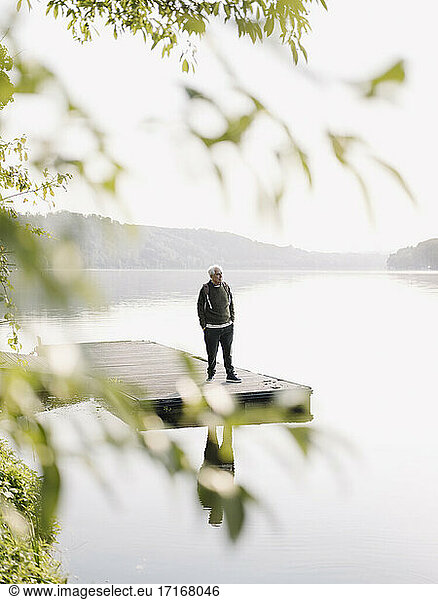 Älterer Mann mit Blick auf den See  während er mit den Händen in den Taschen auf dem Pier steht
