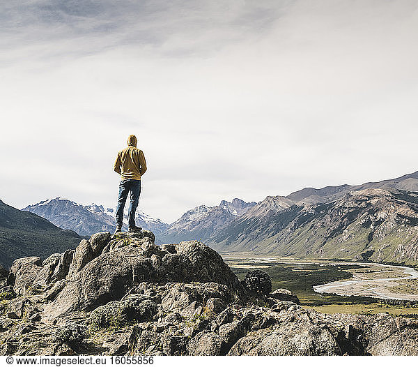 Älterer Mann mit Kapuze  der auf einem Felsen stehend die Berge gegen den Himmel betrachtet  Patagonien  Argentinien