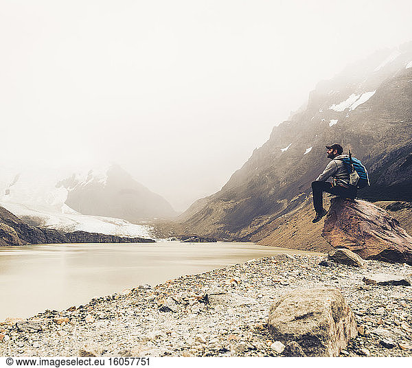 Älterer Mann mit Rucksack  der auf einem Felsen sitzend den See betrachtet  Patagonien  Argentinien