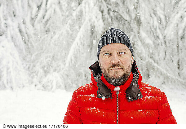 Älterer Mann mit Strickmütze  der im Wald steht und starrt  während es schneit
