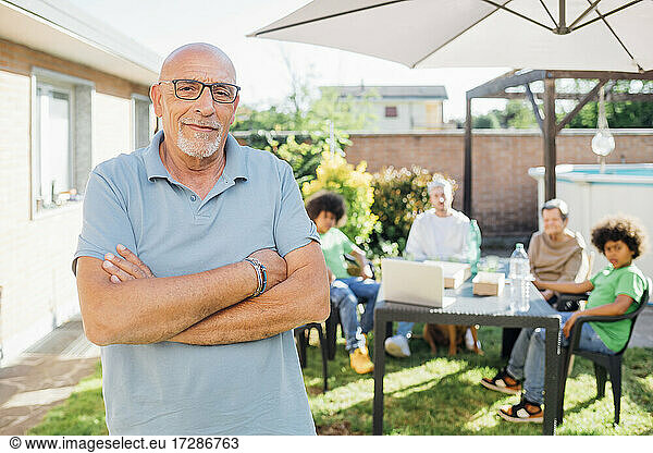 Älterer Mann steht mit verschränkten Armen  während die Familie im Hintergrund im Hinterhof steht