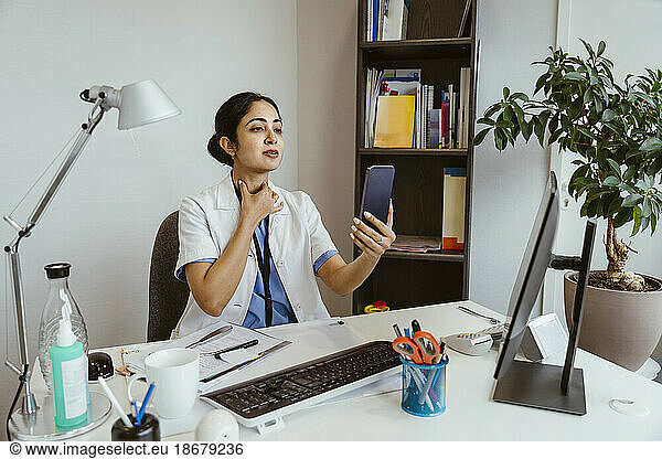 Ärztin bei der Aufklärung eines Patienten per Online-Videoanruf über das Mobiltelefon  während sie am Schreibtisch in der Klinik sitzt
