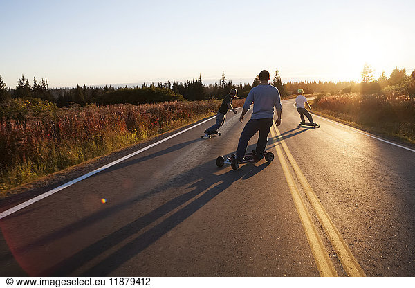 'Three young men skateboarding down a road at sunset; Homer  Alaska  United States of America'
