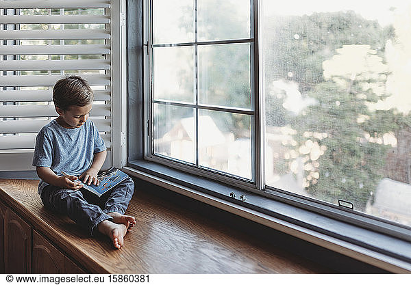 5 yr old boy reading book while sitting on window seat