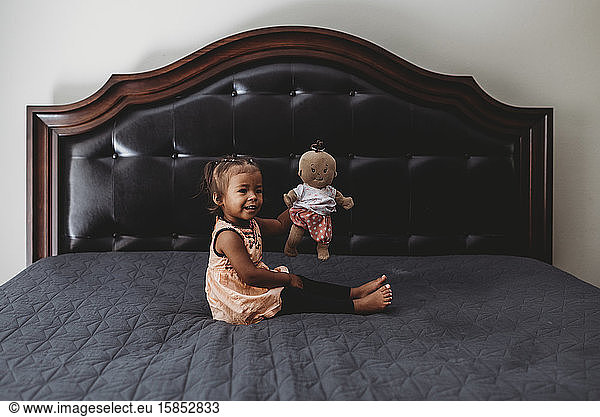 2 yr old multiracial girl playing with doll on parents' bed