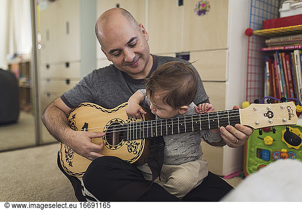 1 yr old playing child's guitar while sitting on bald father's lap