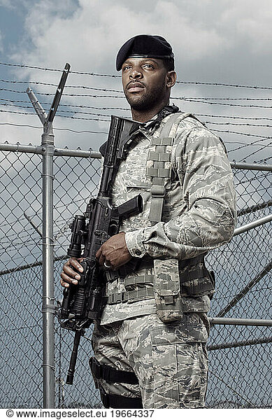 A African American  male  Air Force Security Forces Airman in uniform poses with his M-4 rifle near the a secured fence line with barbed wire.