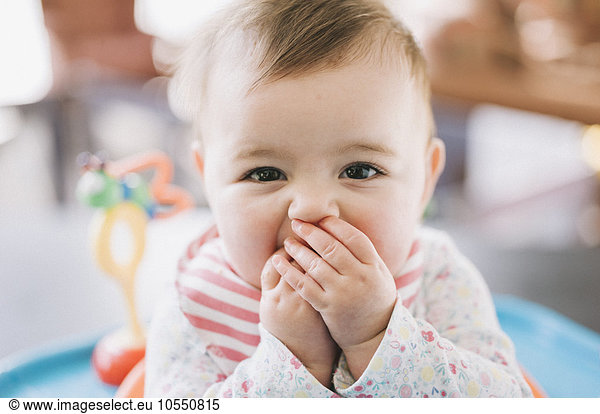 A baby girl with her hands covering her mouth  looking at the camera.