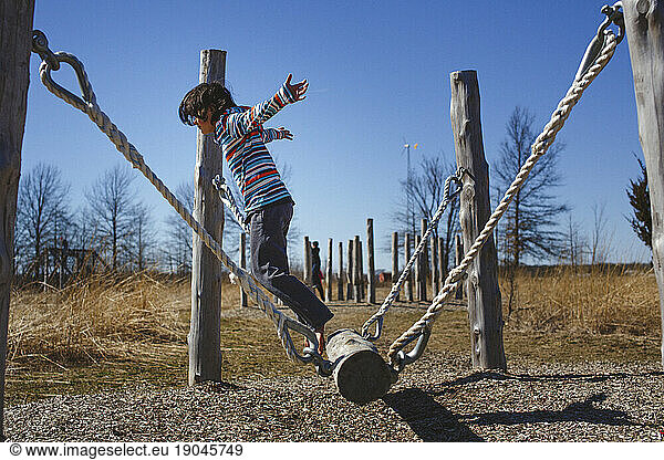 A barefoot child with arms outstretched leaps of a hanging log in park
