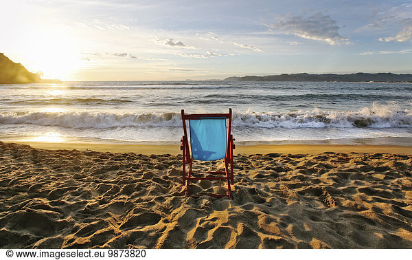A beach chair on the sand and a sunset on the horizon.