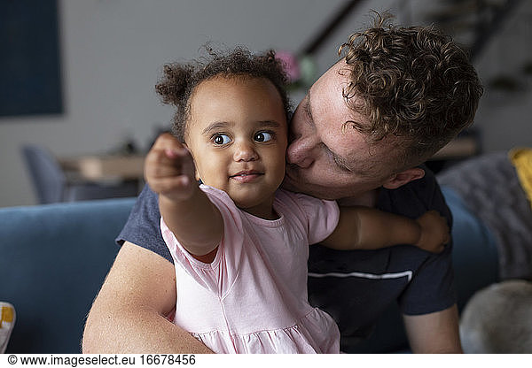 A biracial toddler pointing while her father kisses her cheek