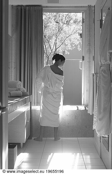 A black and white image of a woman in a bathrobe by a bath tub.