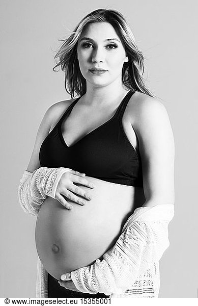 A black and white photo of a young pregnant woman holding her belly in a studio and posing for the camera; Edmonton  Alberta  Canada