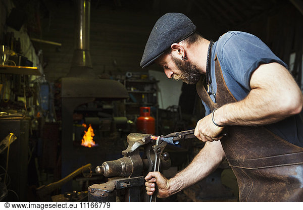 A blacksmith in a leather apron bends a cone of red hot metal in a vice using a wrench and a pair of tongs.