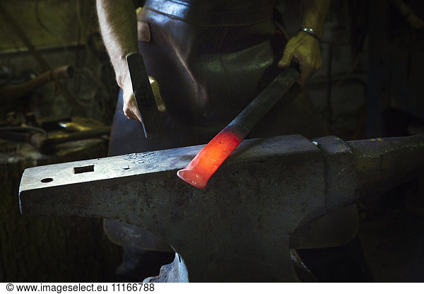 A blacksmith strikes a length of red hot metal on an anvil with a hammer in a workshop.