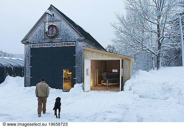 A boat builder  with his dog at his side  walk through the snowy yard after a long winters day in the boat shed.