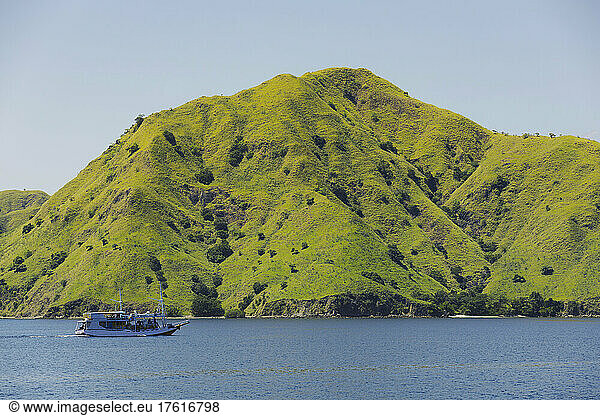 A boat sails along a rugged coastline with landforms covered in green foliage under a blue sky  Komodo National Park; East Nusa Tenggara  Indonesia