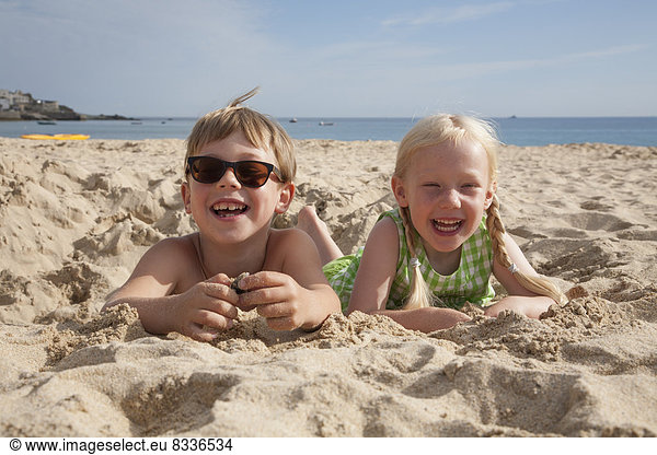 A boy and girl lying on their stomachs on the sand  laughing and looking at the camera.