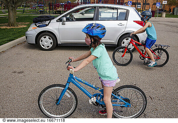 A boy and girl wearing face masks ride bikes together in a parking lot