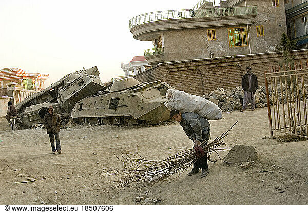 A boy collecting wood and scrap walks in front of destroyed Russian tanks sitting in front of fancy houses in Kabul  Afghanistan