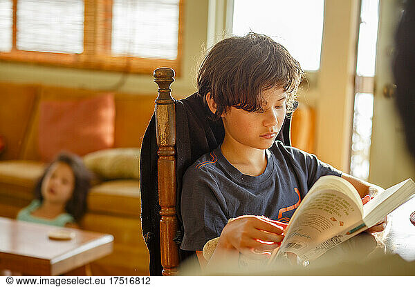 A boy in morning light reads book while sister relaxes behind him