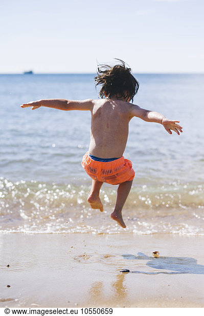 A boy in swimming trunks jumping over waves on the sea shore.