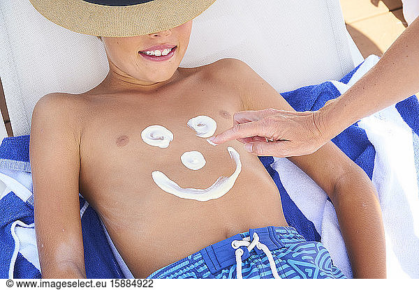 A boy lying on a sun lounger with a woman drawing a face on his chest with sun cream.