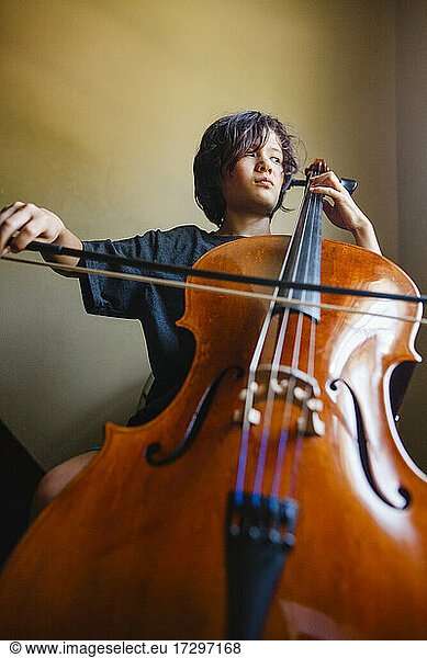 A boy with a serious expression plays cello while looking out window