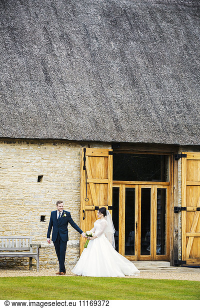 A bride and groom on their wedding day  walking hand in hand from a barn.