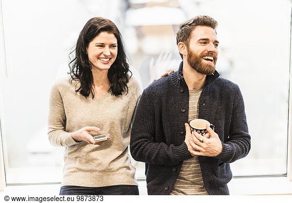 A business woman seated by a window holding a smart phone  talking to a man holding a coffee cup.