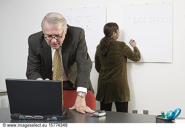 A business women writing on white board with CEO standing by laptop.