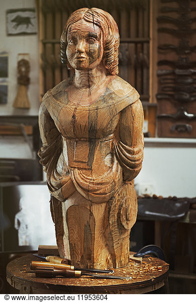 A carved wooden female ship's figurehead on a bench in a workshop with a pattern of wood grain markings