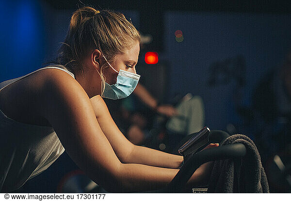 A Caucasian female pedals in an indoor cycling studio with a mask.