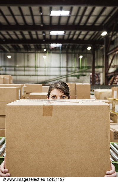 A Caucasian female warehouse worker behind a cardboard box in a distribution warehouse.