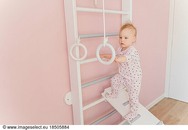 A Caucasian girl in soft pink pajamas does sport at gymnastic rings