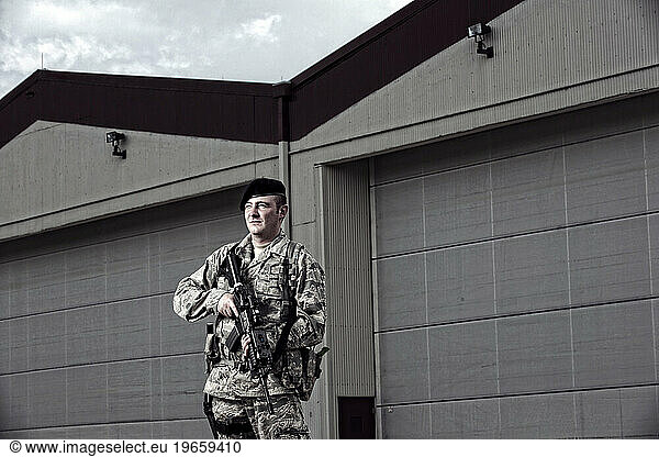 A Caucasian  male  Air Force Security Forces Airman in uniform poses with his M-4 rifle near an airplane hanger on the flight line.